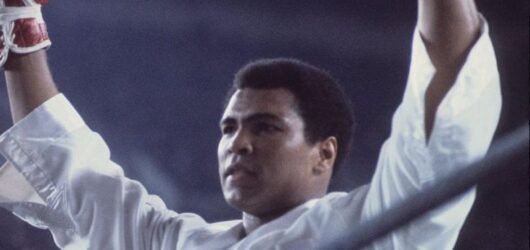 Muhammad Ali raising both gloved hands in victory inside the boxing ring.