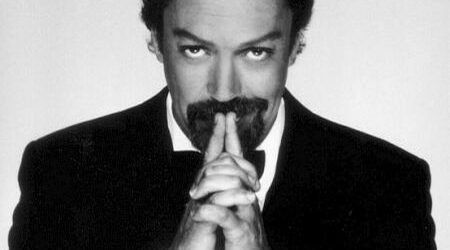 Tim Curry in a black tuxedo with his hands clasped