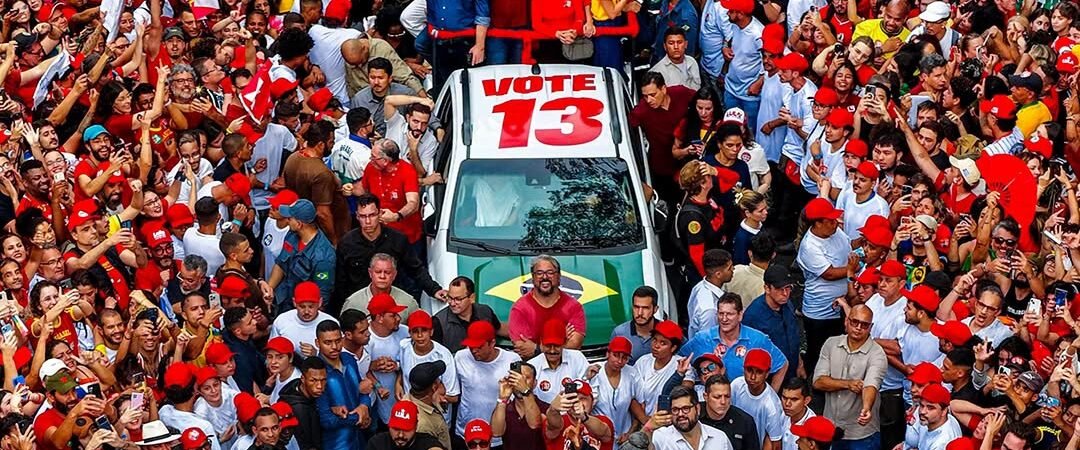 Luiz Inácio Lula da Silva stands on a campaign vehicle surrounded by cheering supporters waving Brazilian flags.