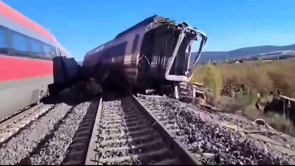Two damaged high-speed trains sit on railway tracks after a collision, with a severely damaged carriage visible beside the tracks.